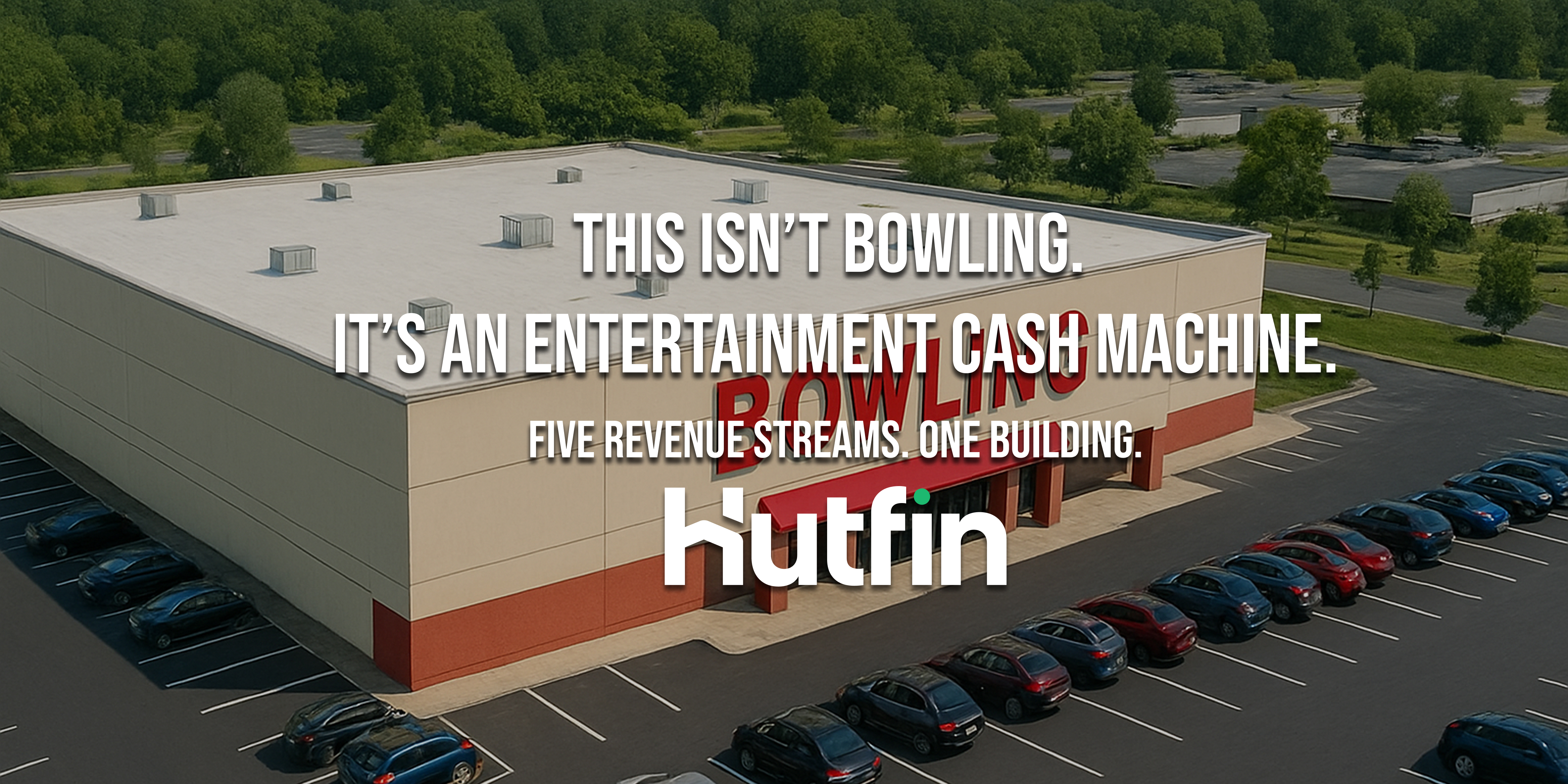 A large, modern single-story commercial building with a black rooftop, surrounded by an expansive parking lot and lush green landscape, photographed from an elevated angle on a clear day.